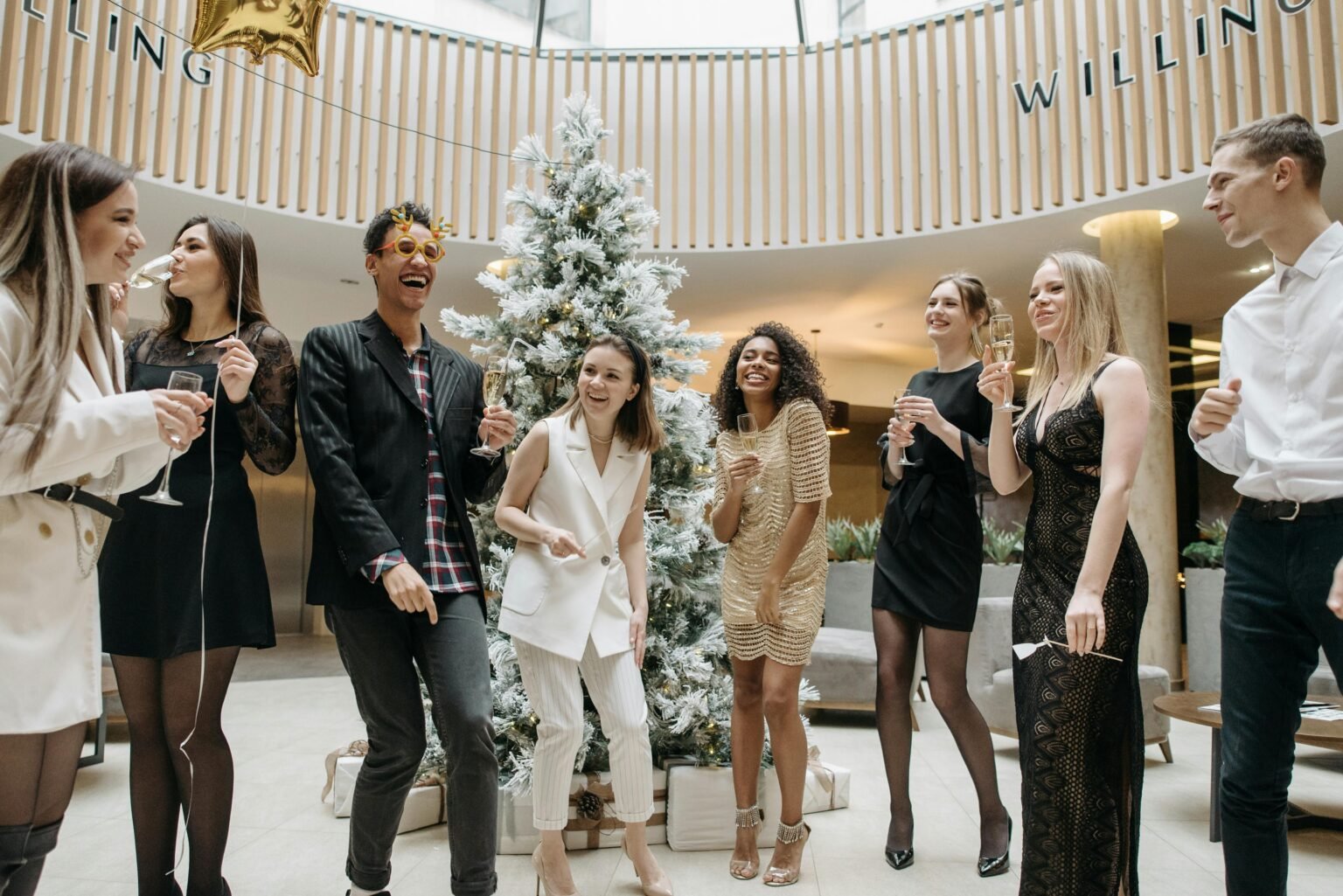 Group of friends celebrating indoors with drinks at a Christmas party by a decorated tree.